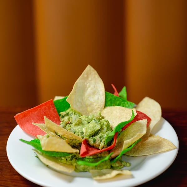 A white plate holds a mound of guacamole surrounded by red, green, and white tortilla chips.