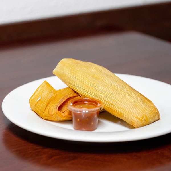 Two golden tamales and a small cup of red salsa on a white plate from Taqueria Taxco.