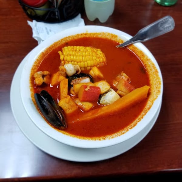 A vibrant red seafood soup with corn on the cob, mussels, and vegetables in a white bowl.