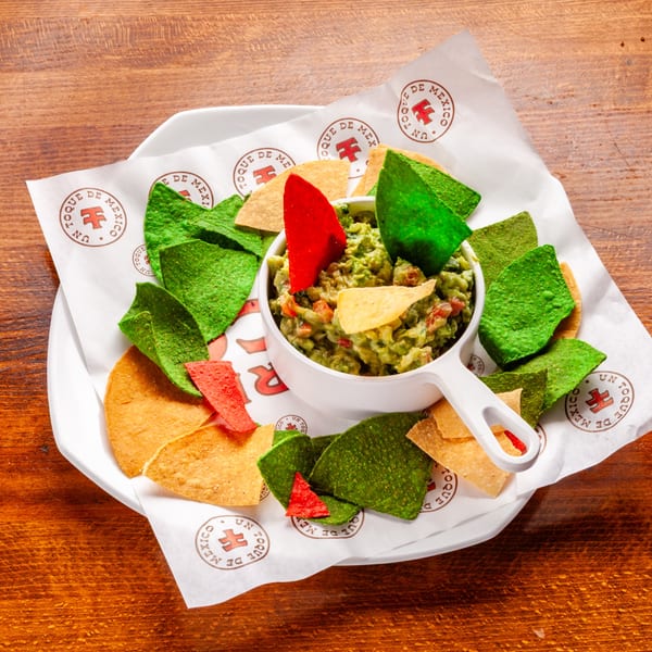 Guacamole with colorful tortilla chips on a white plate with branded paper, on a wooden table.