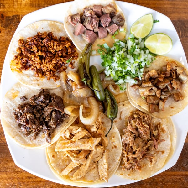 A white plate holds seven Taqueria Taxco tacos with various meats, onions, cilantro, limes, and peppers.