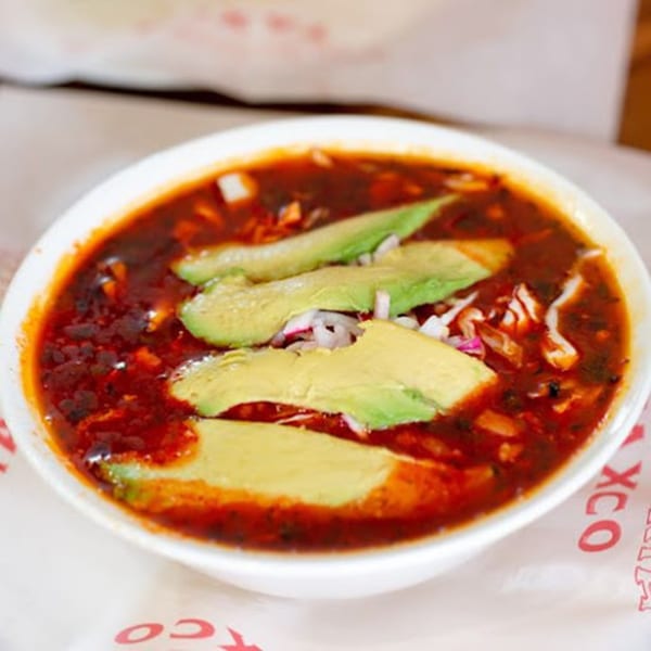 A bowl of red Mexican soup with avocado slices, onions, and cabbage on a branded paper.