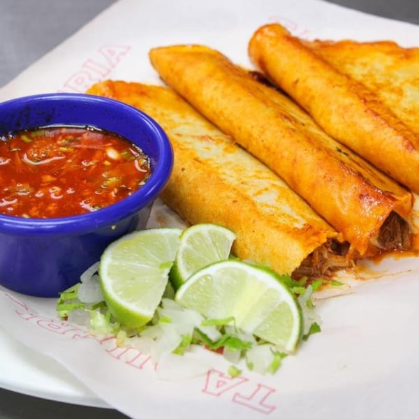 Four golden-brown Taqueria Taxco flautas with a side of red salsa, lime wedges, and chopped lettuce.