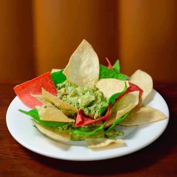 A white plate holds a mound of guacamole surrounded by red, green, and white tortilla chips.