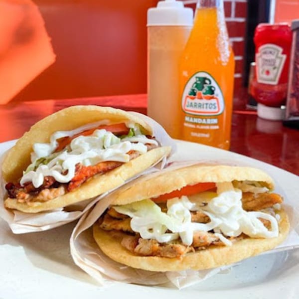 Two Mexican gorditas with meat, lettuce, tomato, and crema, with a Jarritos soda in the background.