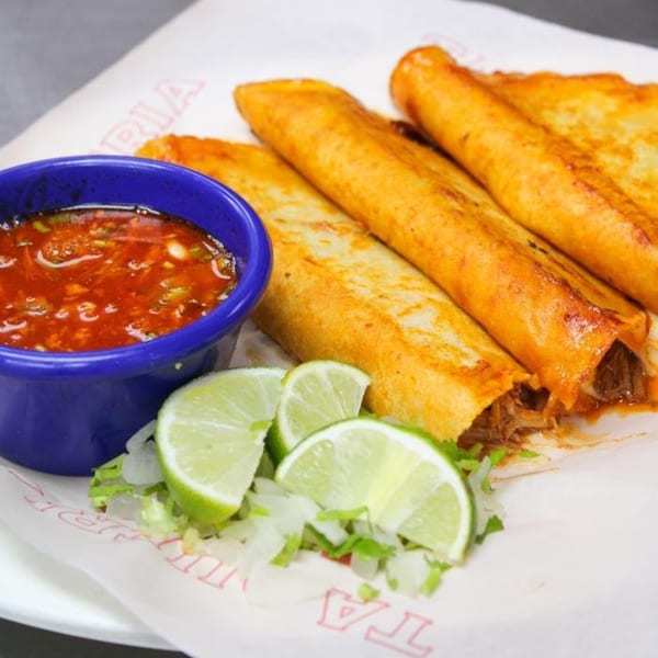 Four golden-brown Taqueria Taxco flautas with a side of red salsa, lime wedges, and chopped lettuce.