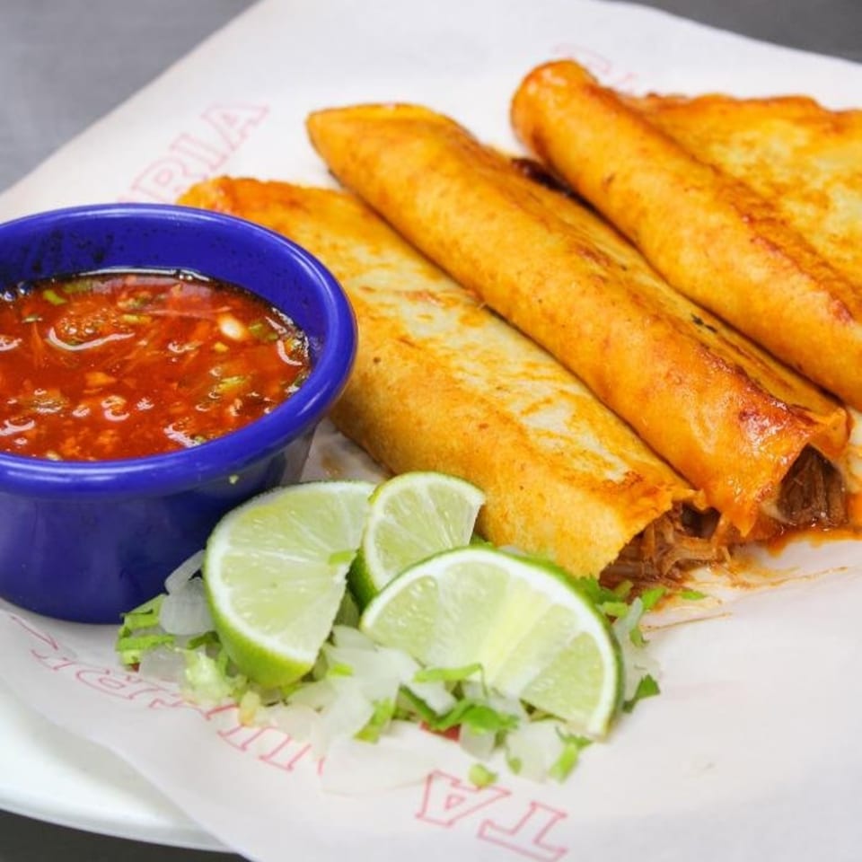 Four golden-brown Taqueria Taxco flautas with a side of red salsa, lime wedges, and chopped lettuce.