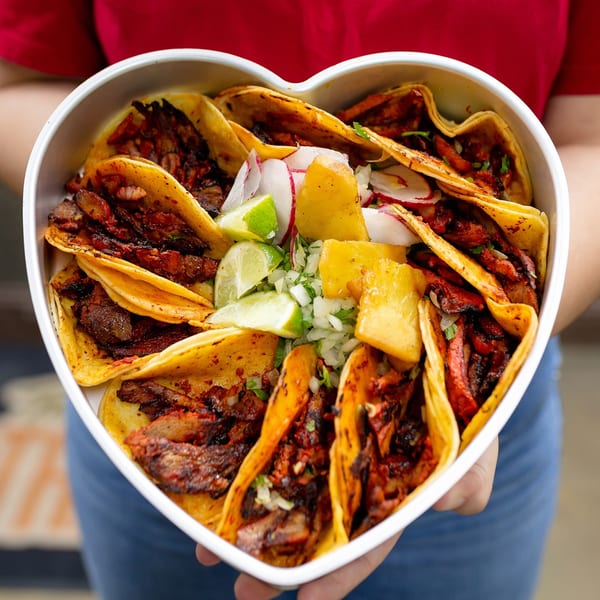 Heart-shaped tray filled with Taqueria Taxco tacos, garnished with lime, radish, pineapple, and onions.